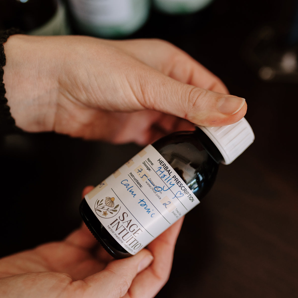 Hands holding a Sage Intuition custom herbal remedy bottle with a handwritten prescription label and white cap.
