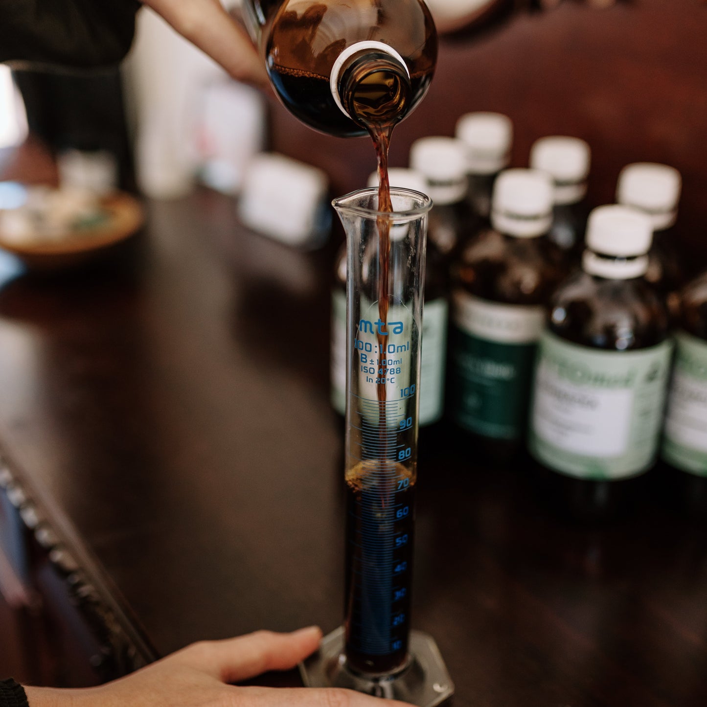 Person pouring a dark liquid from a bottle into a glass container on a wooden surface with other bottles in the background.