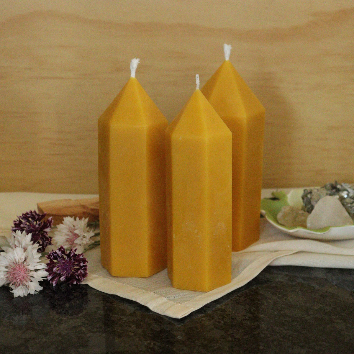 Three tall, yellow beeswax candles on a dark surface with a wooden background.