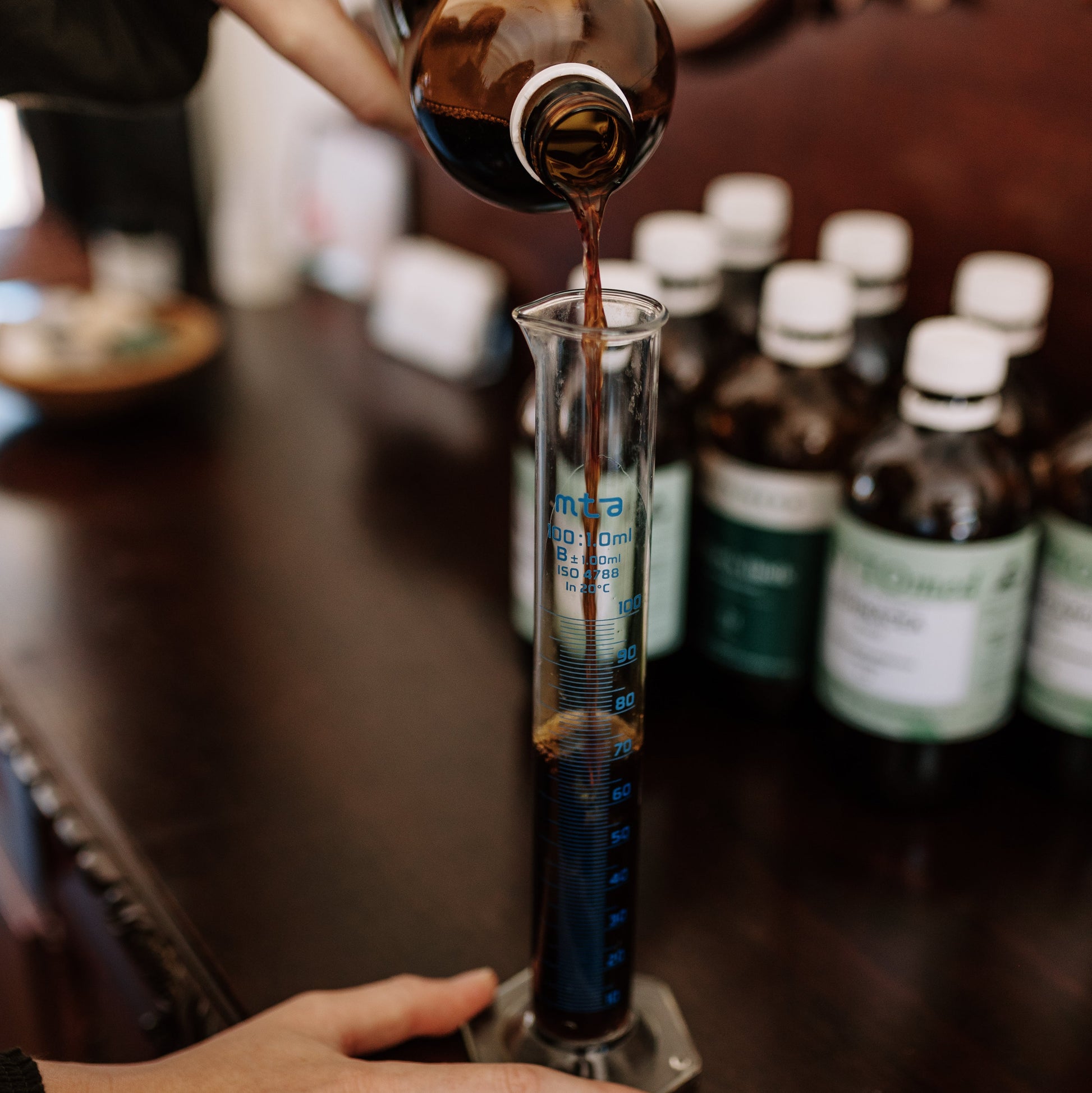Person pouring a dark liquid from a bottle into a glass container on a wooden surface with other bottles in the background.