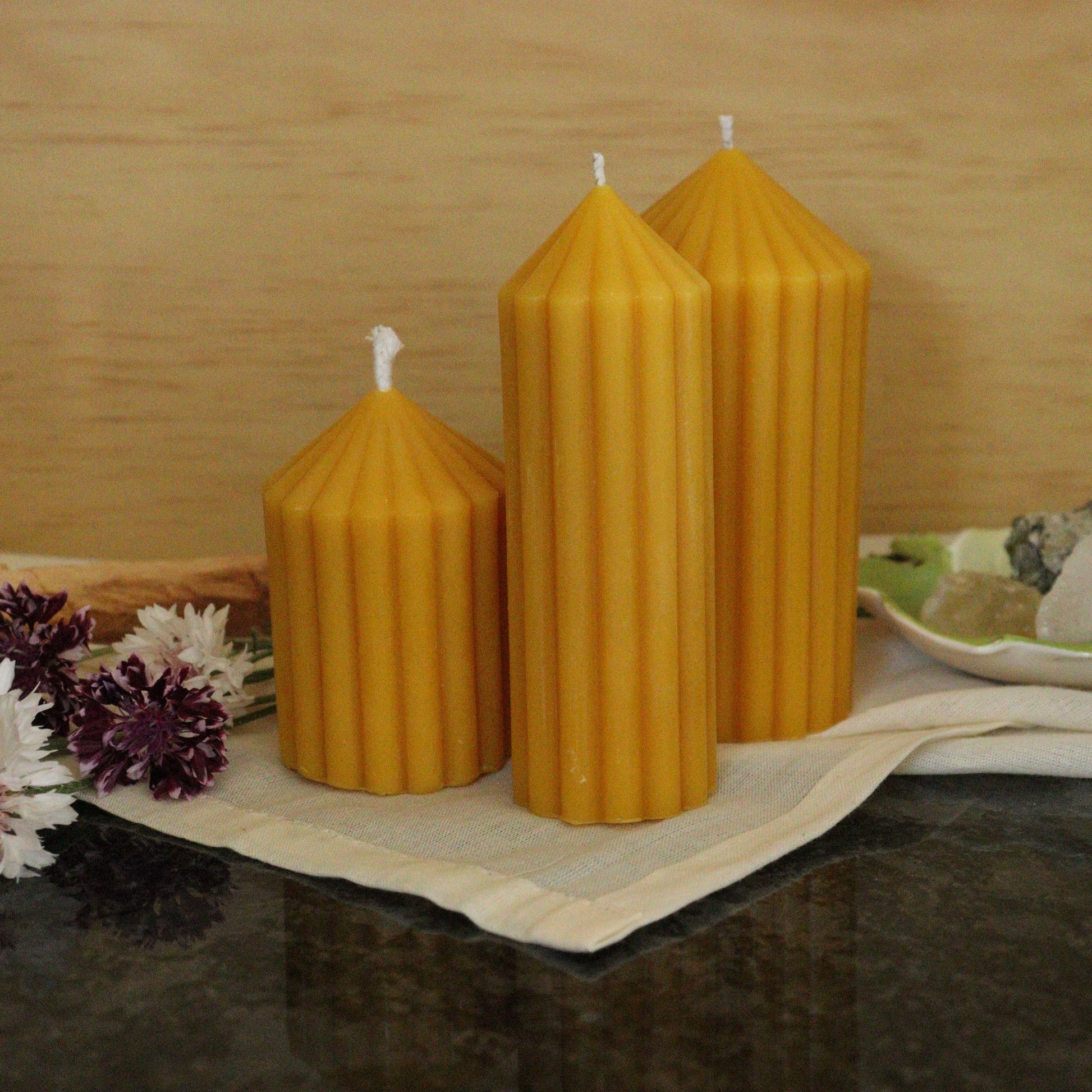 Three beeswax candles on a cloth with flowers and a wooden box in the background.