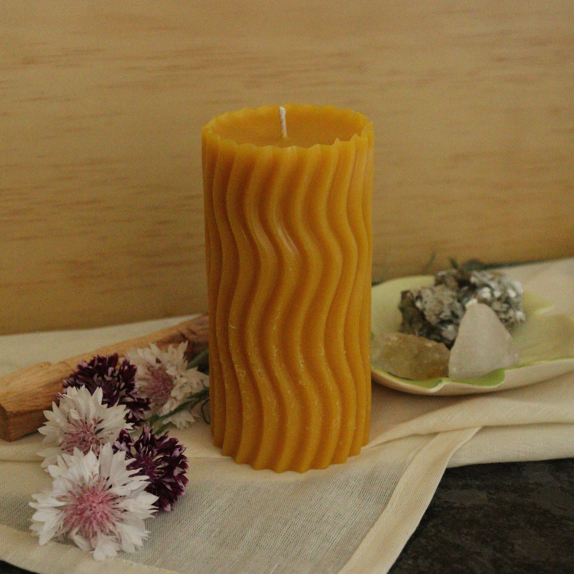 Wavy-textured yellow candle on a cloth with flowers and a rock in the background